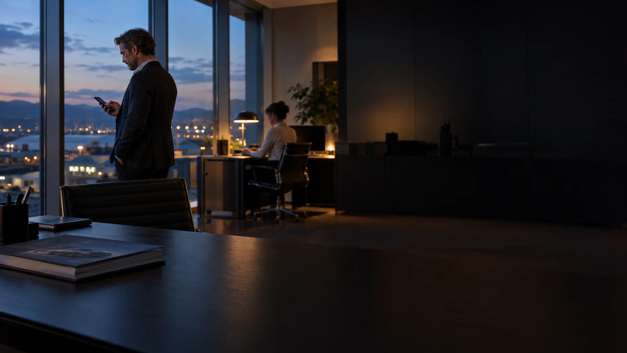 Regional manager reading a phone by an office window at dusk while a colleague works at a desk